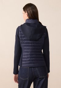Námornícky puffer vest s rebrovanou textúrou, pripevnenou kapucňou a hladkými rukávmi; priliehajúci strih s kontrastnými látkami a stehmi.