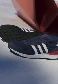 Zapatilla Adidas azul oscuro con rayas blancas y detalle rojo en el talón, mostrada con la suela hacia arriba, sobre una superficie de hormigón clara junto a una escultura geométrica de color óxido.