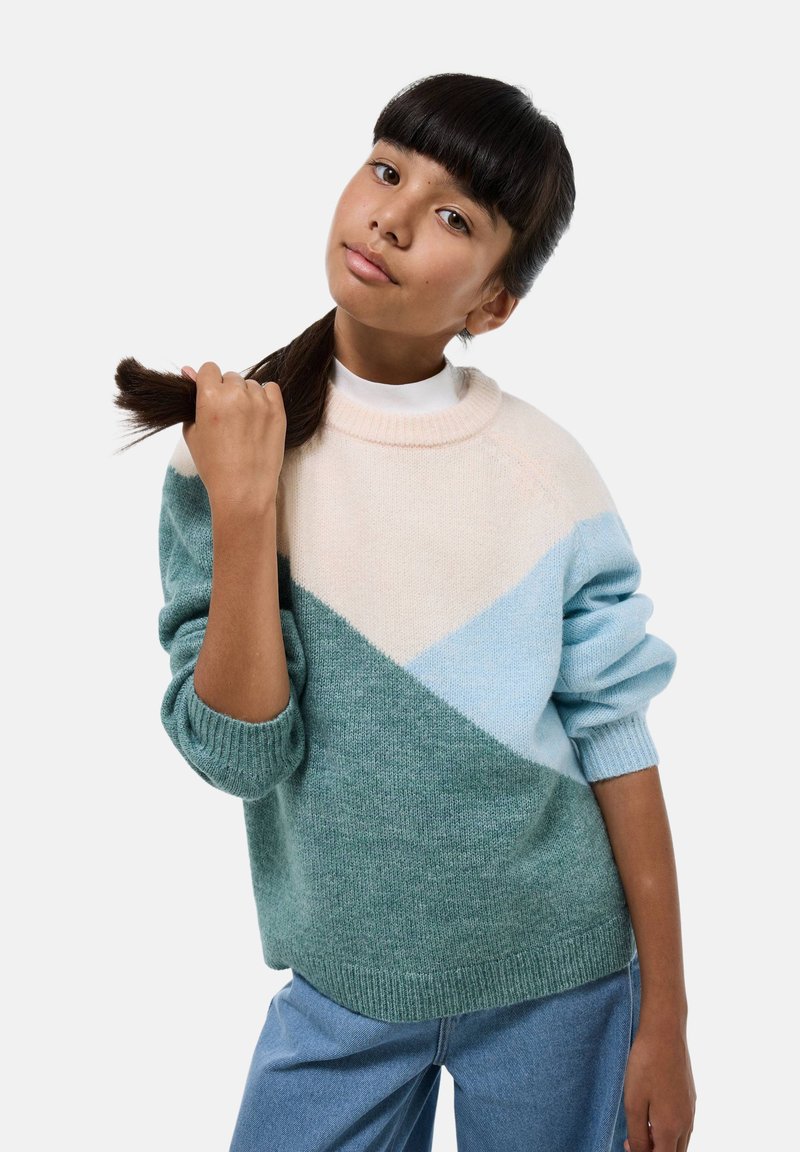 Pull en tricot avec un design en blocs de couleur dans des teintes de vert menthe, bleu pâle et crème. Il présente un col rond et des poignets côtelés, porté avec un jean décontracté.