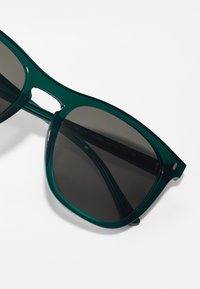 Ochelari de soare din plastic verde, cu lentile întunecate, design unghiular și accente tăiate. Prezintă o formă elegantă și o textură netedă.