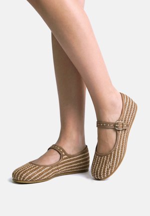 Voeten met bruine en witte geweven Mary Jane platte schoenen met gespsluiting, geposeerd tegen een effen witte achtergrond.