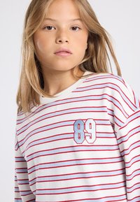 Young girl with straight blonde hair wearing a white and red striped shirt with "89" embroidered on the chest.