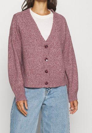 Kobieta w mauve kardiganie zapinanym na guziki na białą koszulę, zestawiona z jasnoniebieskimi jeansami z wysokim stanem, stojąca na jednolitym tle.