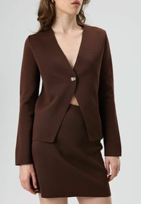 Bruine blazer met een diepe V-hals en een enkele knoopsluiting, gecombineerd met een bijpassende rok; gestructureerde stof; getailleerde pasvorm; minimale details.
