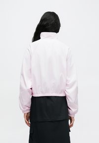 Ροζ ανοιχτό ροζ σακάκι με ελαστική μέση και μανσέτες, με ψηλό γιακά. Το ύφασμα φαίνεται λείο και αδιάβροχο, σχεδιασμένο με κομψό cropped στυλ.