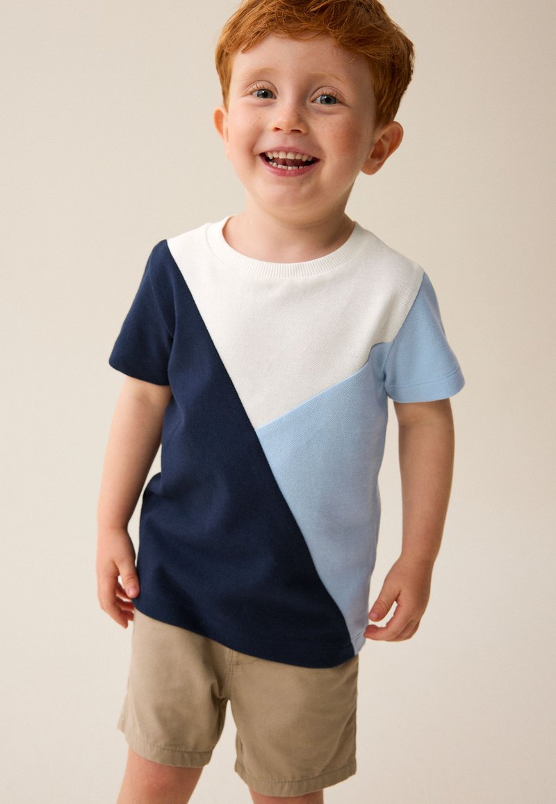 Rothaariger Junge lächelt, trägt ein kurzärmeliges Farbblockshirt in Marineblau, Weiß und Hellblau mit beigen Shorts vor einfarbigem Hintergrund.