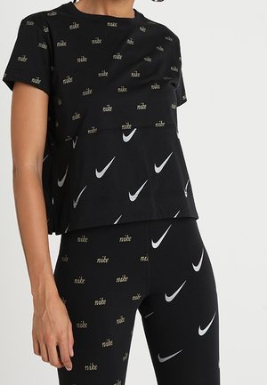Osoba nosząca czarny strój Nike z powtarzającym się małym złotym tekstem "nike" na górze i legginsach oraz białymi logo Nike swoosh na górze i legginsach.
