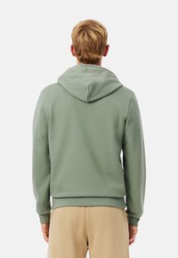 Sudadera con capucha verde hecha de tejido suave, cuenta con capucha con cordón y puños acanalados. Textura lisa con un diseño sencillo, combinada con pantalones beige claro.