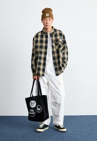 Camisa de franela de cuadros en beige y negro, camiseta blanca, pantalones cargo blancos con bolsillos, bolsa tote negra con gráficos y zapatillas verdes.