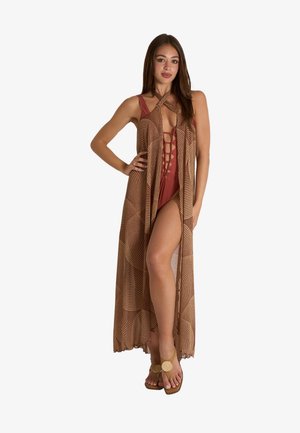 Donna con costume intero color ruggine con allacciatura frontale, indossa un lungo copricostume marrone con fantasia e sandali beige, posa con la mano sul fianco.