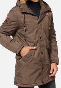 Bruine geïsoleerde parka met een capuchon, gebaseerd op nepbont voering, verstelbare trekkoorden, patchdetails en voorkezen met drukknoopsluitingen.