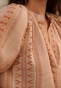 Blusa color pesca con motivi a righe verticali e geometrici. Tessuto morbido con dettaglio delle spalle arricciato e scollatura a V. Texture sottili.