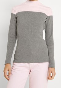 Nainen, joka on pukeutunut ribbimäiseen harmaaseen neuleeseen, jossa on vaaleanpunainen yoke ja vaaleanpunaiset housut, seisoo yksinkertaista vaaleaa taustaa vasten.