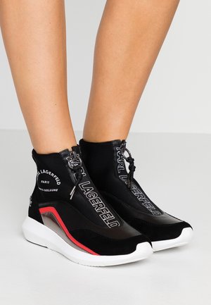 Baskets montantes noires avec semelles blanches, accents de rayures rouges, fermetures éclairs à l'avant et marquage "KARL LAGERFELD" portées sur des jambes nues.