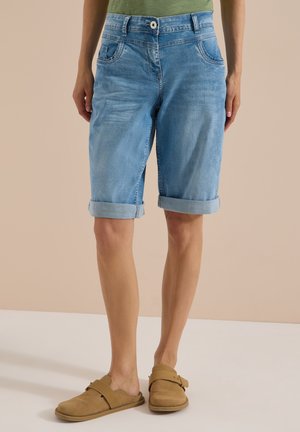 Personne portant un short en jean bleu clair à revers, une chemise verte et des chaussures beige sans lacets, debout devant un fond beige.