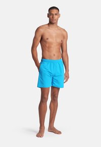 Modelo masculino de pie descalzo con las manos en los bolsillos, vistiendo unos bañadores de color azul brillante contra un fondo blanco liso.