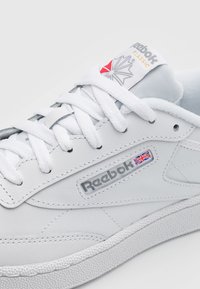 Baskets blanches en cuir avec semelle en caoutchouc texturée, arborant un logo Reebok gris, un patch logo rouge et gris, et des lacets plats blancs.