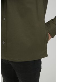 Camisa verde oliva con cierre de botones, con botones a presión de metal y un corte relajado. La textura parece suave con un detallado mínimo.