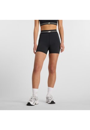 Femme portant un short de sport taille haute noir et un crop top assorti, des chaussettes blanches et des baskets New Balance blanches, debout sur un fond gris.