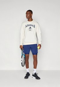 Adidase kreemikas pusa tumesinise kirjaga, tumesinised lühikesed püksid, mustad tossud, tennisereketi ja palliga, ühtlasel valgel taustal.