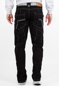 I pantaloni cargo neri presentano cuciture bianche, molteplici tasche e un design aderente. Abbinati a scarpe da ginnastica bianche, mostrando il dettaglio del tallone.