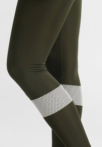 Tmavě zelené legíny s hladkou texturou, které mají bílou síťovanou vložku s kruhovými vzory kolem oblasti středního lýtka.