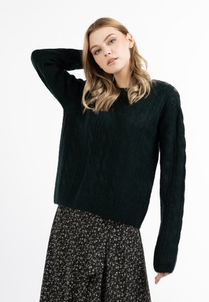 Blondynka ubrana w czarny sweter z warkoczowym splotem i czarną kwiecistą spódnicę, pozująca z jedną ręką za głową na białym tle.