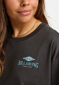 Černé bavlněné tričko s tyrkysovým a růžovým logem Billabong, kulatým výstřihem a krátkými rukávy. Hladká textilie, volný střih.
