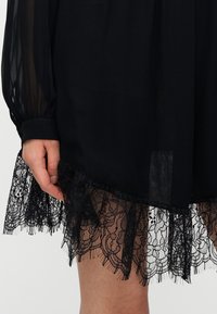 Μαύρο φόρεμα με διαφανή μανίκια, που διαθέτει μαύρη δαντέλα με κυματωτό τελείωμα. Η δαντέλα έχει floral μοτίβα και υφή.