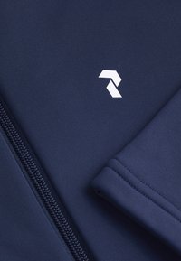 Marineblå sportsjakke med struktureret stof, der har en lynlås og et præget hvidt logo øverst til venstre.