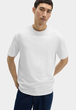Mężczyzna z krótkimi ciemnymi włosami ubrany w zwykły biały T-shirt i ciemne spodnie, stoi z rękami za plecami na jasnym tle.