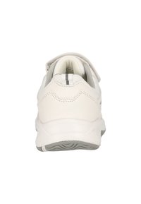 Chaussure de sport blanche avec une tige texturée, un col rembourré et une sangle ajustable. Dotée d'une semelle grise avec un motif ondulé et des accents argentés.