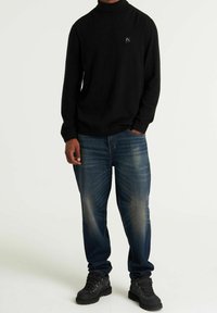 Schwarzer Rollkragenpullover mit kleinem Logo, kombiniert mit blauen Jeans aus Denim. Die Schuhe sind dunkel und robust, geeignet für den Einsatz im Freien.