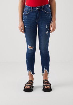 Mørkeblå skinny jeans med mellemhøj talje, med revnede detaljer på låret og frynsede kanter, båret med sorte remsandaletter og rød top.