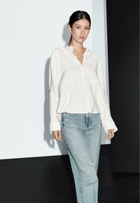 Vit skjorta med rund krage, oversized passform och långa ärmar, som kombineras med ljusblå raka jeans. Slät tygtextur.
