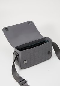 Šedá texturovaná crossbody taška s geometrickým vzorem, vybavená klopňovým uzavřením, kovovým hardwarovým prvkem a nastavitelným popruhem. Uvnitř je podšívka v sytě modré barvě.
