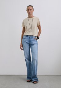 Camiseta de algodón beige, de corte relajado, combinada con jeans acampanados de color azul claro. Collar largo negro con acento circular, pulsera negra y sandalias.