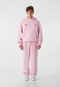 Svetlo roza kapuca in ujemajoče se trenirke iz mehkega materiala. Kapuca ima sprednji žep in vezenje; sproščen kroj primeren za vsakodnevno nošenje.