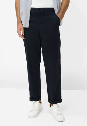 Mann trägt hochgekrempelte schwarze Hose, weiße Turnschuhe, ein weißes T-Shirt und ein hellgraues offenes Hemd, steht mit einer Hand in der Tasche.