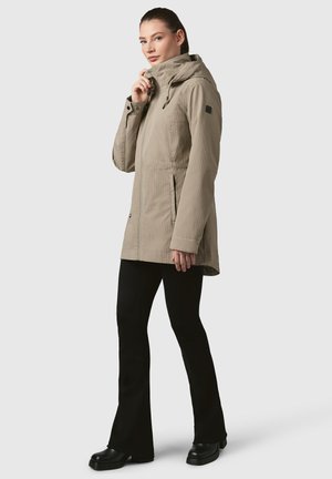 Veste imperméable beige avec capuche, poches latérales et fermeture éclair. Tissu lisse et léger avec une texture subtile. Associée à un pantalon noir.