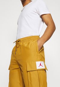 Pracovní kalhoty z hořčicově žluté látky, s elastickým pasem, bočními kapsami a bílou etiketu s červeným logem Jordan.