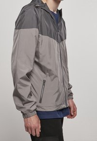 Man som bär en tvåtonad grå zip-up jacka med dragkedjeförsedd sidokok, stående mot en enfärgad ljusgrå bakgrund.