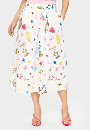 Vrouw die een witte midi-rok draagt met kleurrijke abstracte vormen en rode, blauwe, gele en groene patronen, gecombineerd met roze sandalen met bandjes.