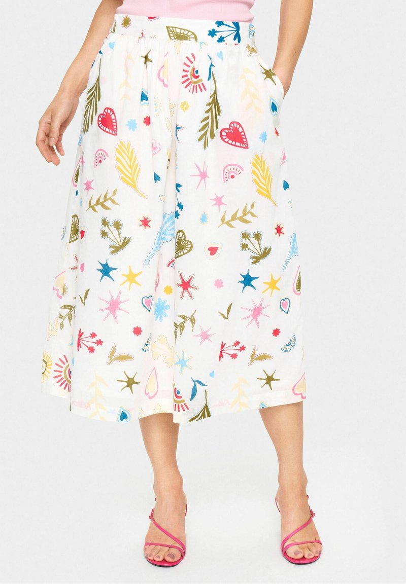 Femme portant une jupe midi blanche avec des formes abstraites colorées et des motifs rouges, bleus, jaunes et verts, associée à des talons roses à brides.