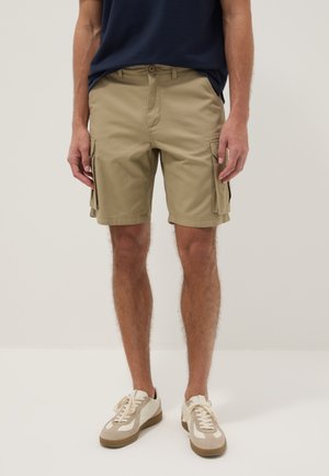 Hombre con pantalones cortos cargo beige, camiseta azul marino y zapatillas blancas, de pie frente a un fondo liso.