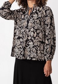 Blusa nera con pattern floreale bianco, maniche lunghe gatherate ai polsini, scollatura a chiave con dettaglio a fiocco e vestibilità morbida.
