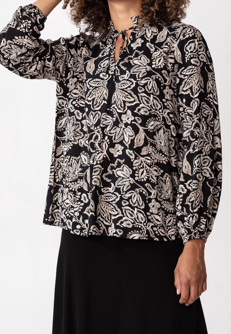 Blusa nera con pattern floreale bianco, maniche lunghe gatherate ai polsini, scollatura a chiave con dettaglio a fiocco e vestibilità morbida.