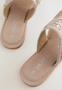 Beige slip-on sandaler med blommönsterbroderi, mjuk mockakänsla och platt sula. Har en rundad form med öppen tå-design.