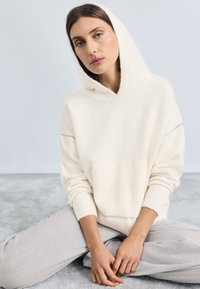 Cream-colored hooded sweatshirt with a textured finish, featuring a front pocket and ribbed cuffs, paired with light gray pants.