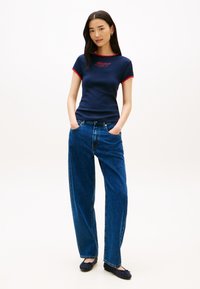 Μπλε ναυτικό T-shirt με κόκκινη διακόσμηση και λογότυπο, συνδυασμένο με φαρδιά μπλε τζιν και σκούρα φλατ παπούτσια. Το μοντέλο στέκεται με τα χέρια στις τσέπες μπροστά σε ουδέτερο φόντο.